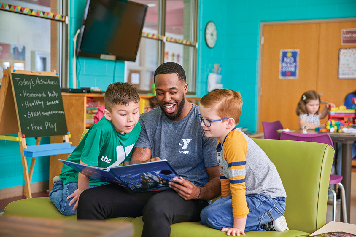Childcare Programs YMCA staff member reading a book to two young boys while seated in a colorful activity room.